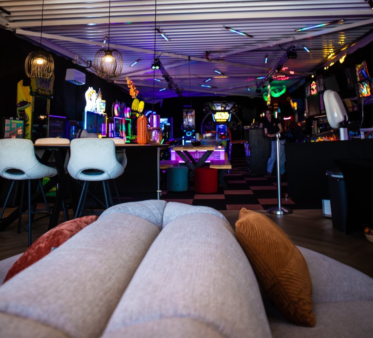 Lounge en chill ruimte in Leusden met een arcadehal met kleurrijke spelautomaten, een grote houten tafel, hanglampen en neonverlichting.