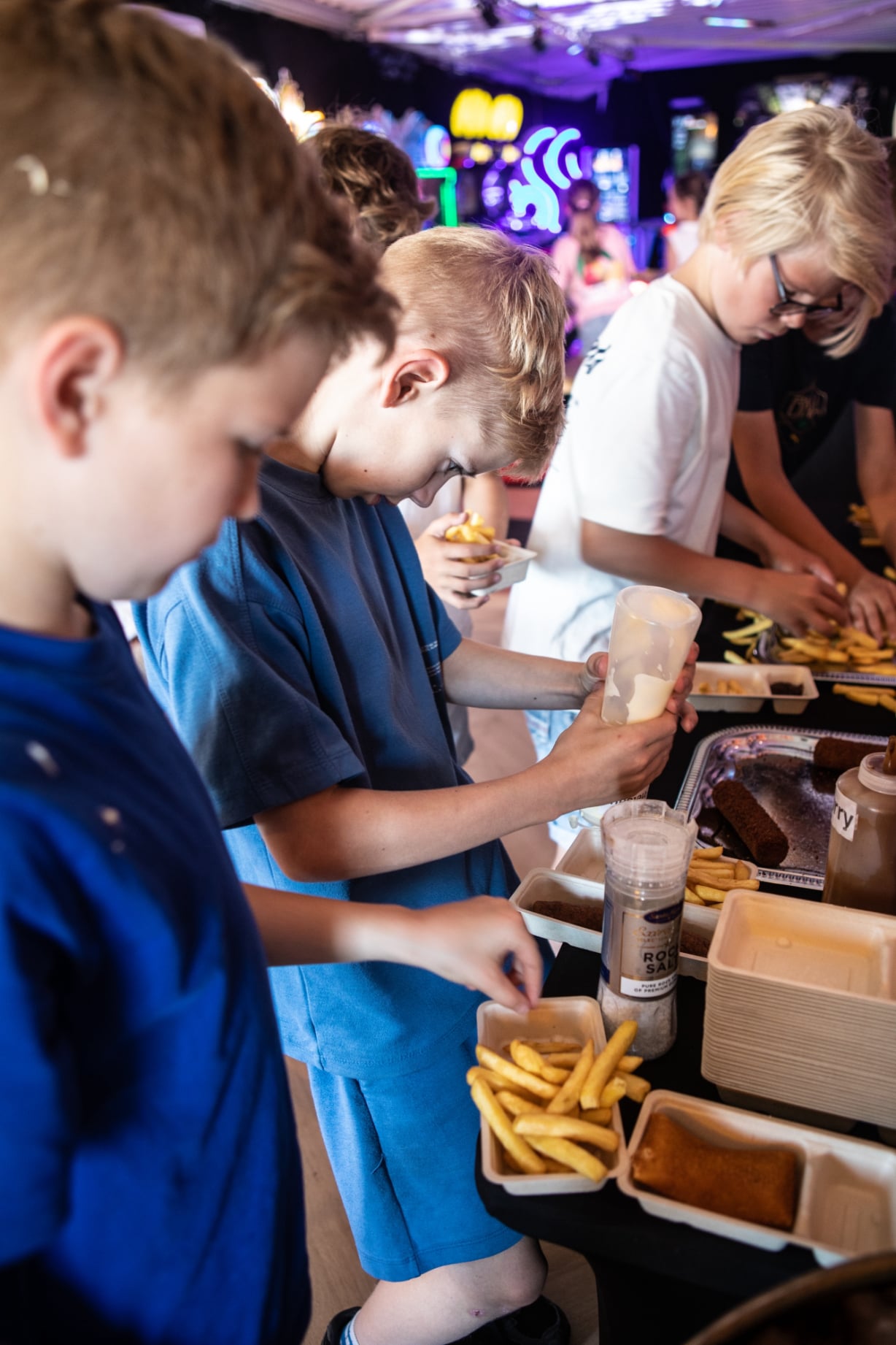Kinderen en volwassenen scheppen friet en snacks op bij een buffet in een sfeervol verlichte gamehal.