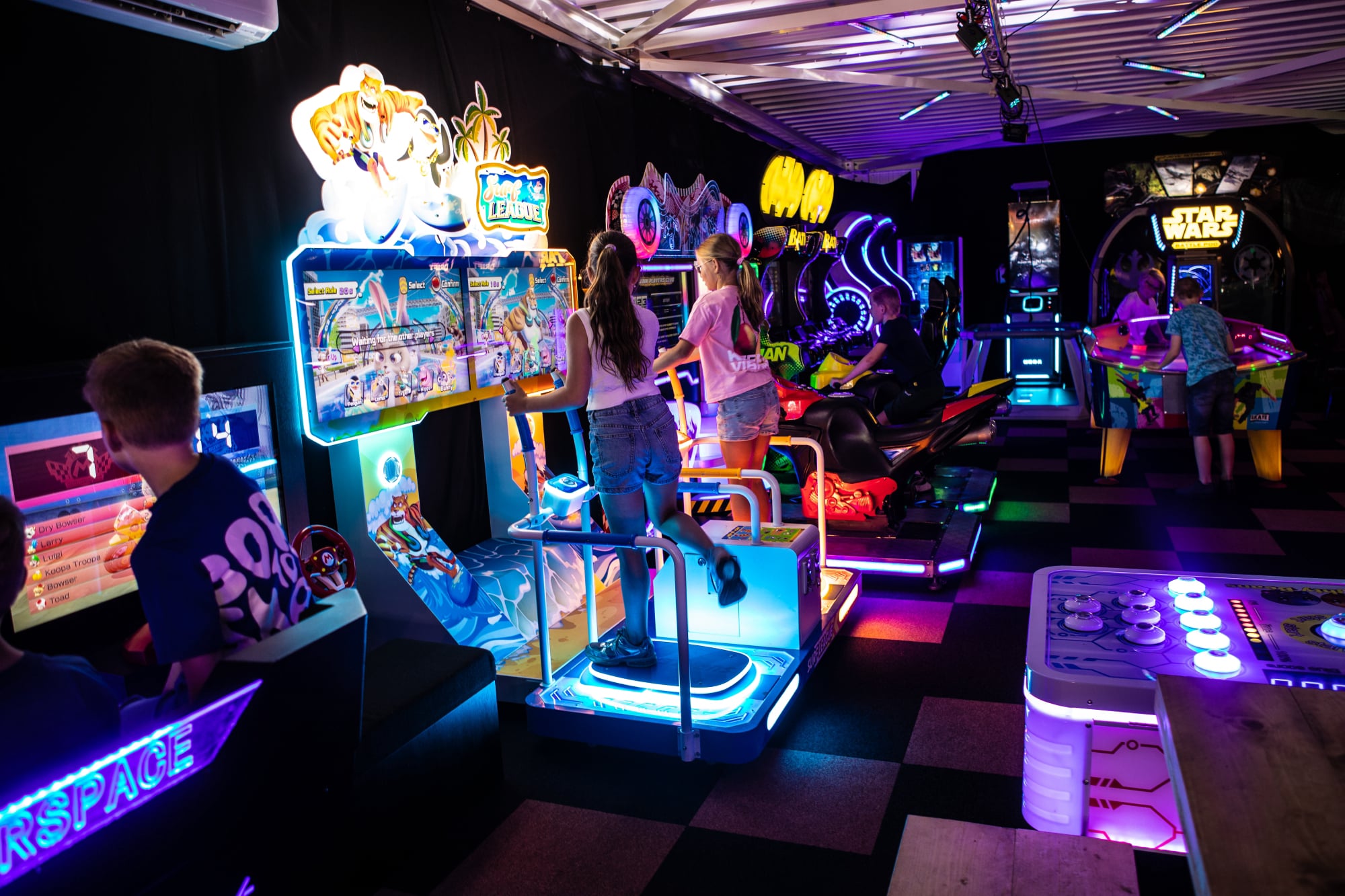 Sfeerimpressie van Game Lounge Leusden met een ruime arcadehal, kleurrijke spelautomaten en bezoekers die samen gamen. De lounge met een grote houten tafel, hanglampen en neonverlichting biedt een gezellige plek om tussen het spelen door te ontspannen.